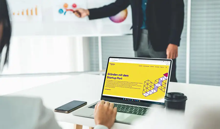 Eine Person arbeitet am Webdesign für eine gelbe Startup-Seite, während eine andere in Hamburg Charts auf einer Tafel dahinter präsentiert.