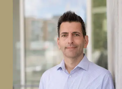 Mann mit kurzen dunklen Haaren in einem hellblauen Hemd, stehend in einem Haus in Hamburg mit unscharfen Fenstern im Hintergrund.